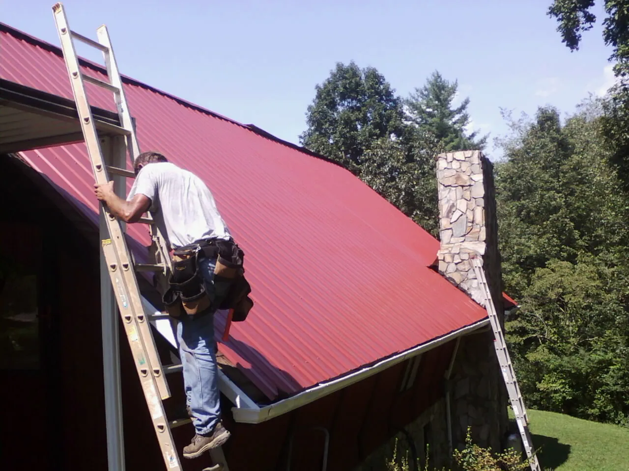 Roofer on a ladder beside a red metal roof and stone chimney — Soffit & Fascia Repair by Roofing Brothers in Talmo